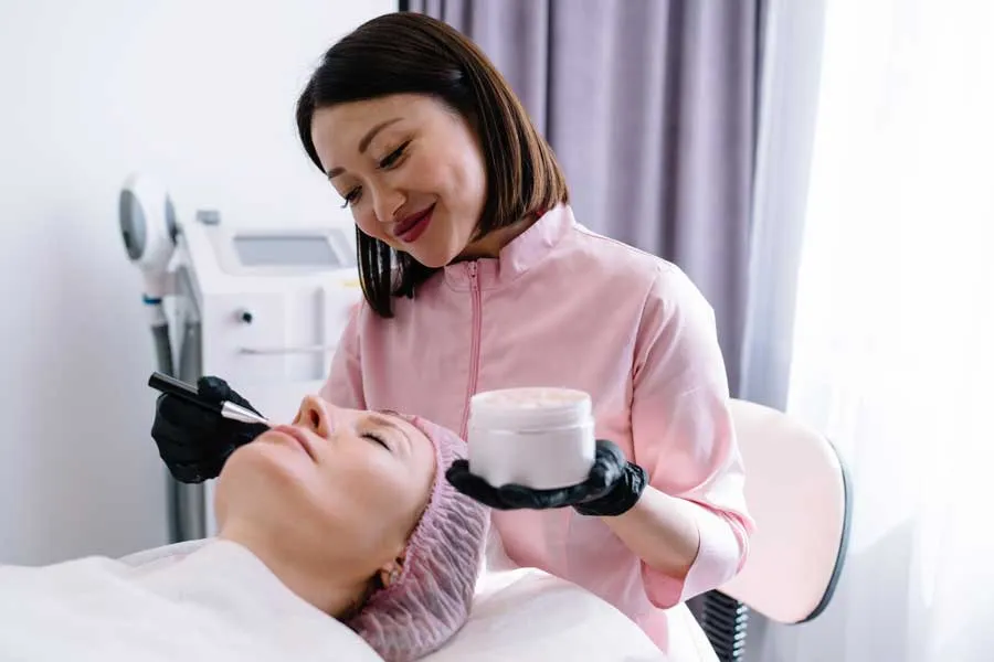 Positive cosmetologist applying cream on client face